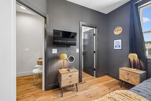 A modern bedroom with a flat-screen TV, a mirror, two bedside tables with lamps, a bed, and a window with curtains.