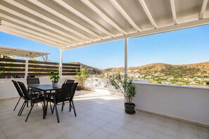 A covered outdoor patio with a table and chairs, potted plants, and a view of a hillside town. - Syros Sun Villa - Terrace Open Views near Airport (Manna)