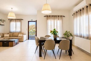 A modern living room with a sofa, dining table, chairs, and a view through a door to an outdoor balcony. - Syros Sun Villa - Terrace Open Views near Airport (Manna)