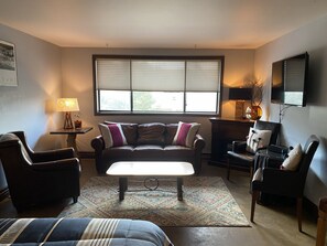 A living room with a sofa, two armchairs, a coffee table, a TV, and a window with blinds. - Perfect Frisco Location: Cozy Studio Steps from Main Street (Frisco)