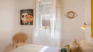 A bedroom with a bed, a chair, a painting, a window with curtains, and a wall-mounted lamp. - Casa François – Appartement rénové à 100 m de la plage de Calvi (Calvi)