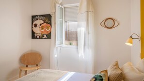 Ein Schlafzimmer mit einem Bett, einem Sessel, einem Gemälde, einem Fenster mit Vorhängen und einer Wandleuchte.