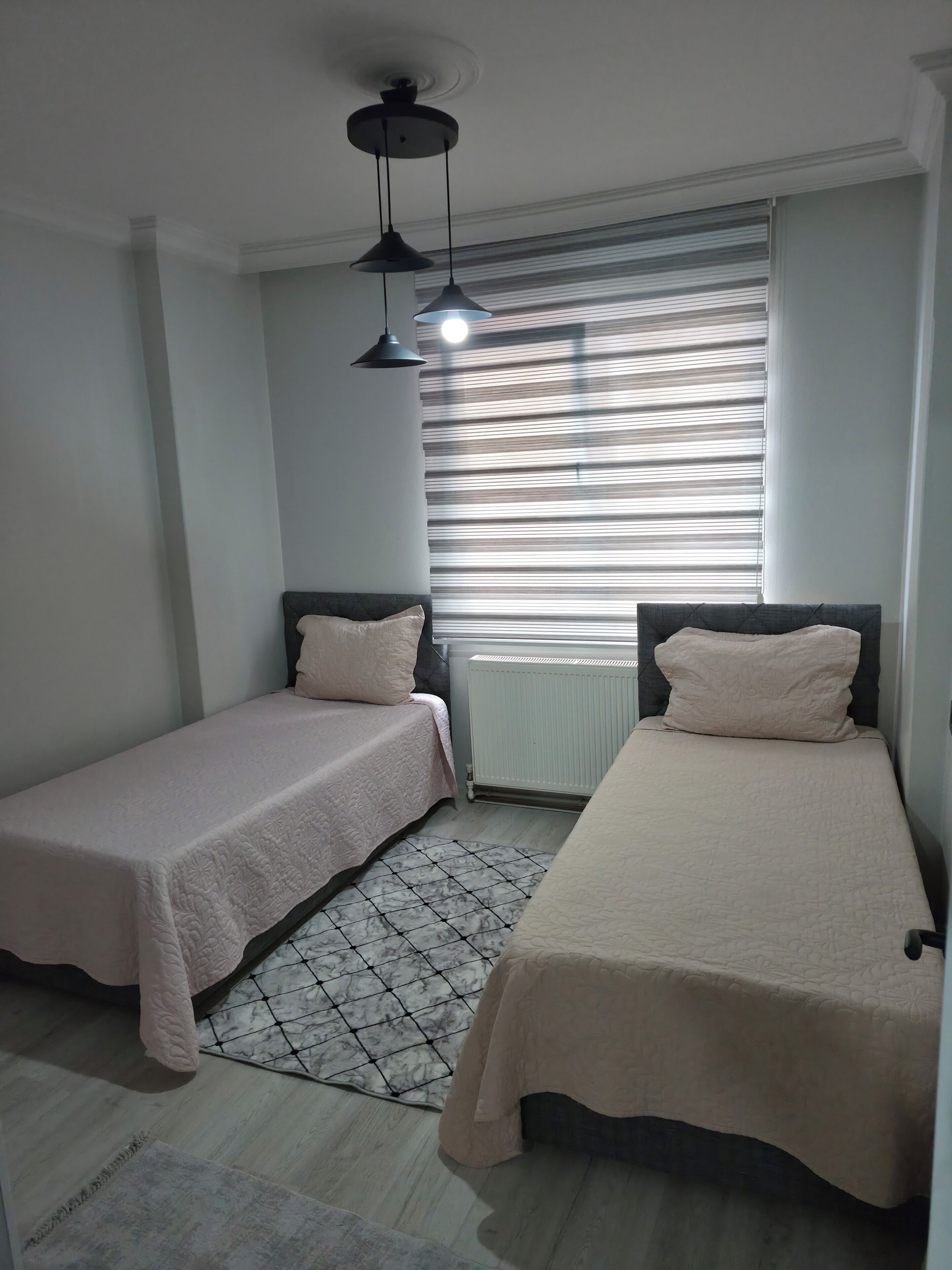 A bedroom with two single beds, a radiator, a window with blinds, and a ceiling fan.