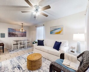 Un salon doté d’un ventilateur de plafond, d’un canapé blanc, d’une table à manger avec des chaises et d’un pouf en osier.