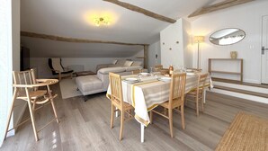 Uma área de refeições com mesa posta para quatro pessoas, cadeiras de madeira, um sofá e uma televisão ao fundo.