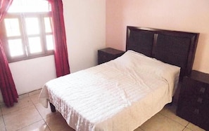 A single bed with a white bedspread in a room with a window and a nightstand.