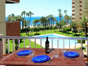 Une table en bois, avec des assiettes bleues, une bouteille et des verres, donnant sur une piscine et des palmiers.