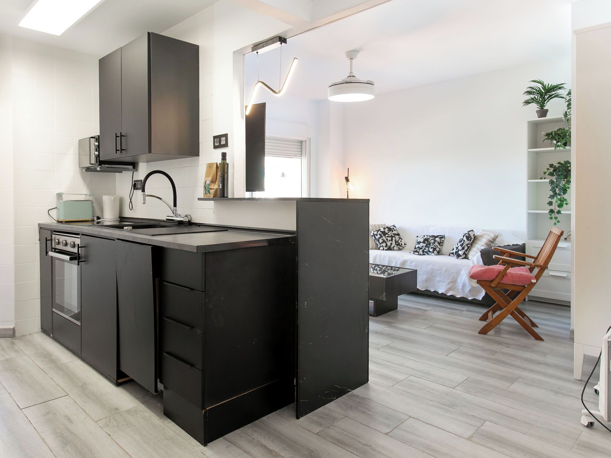 A modern kitchen with a central island, a dining area with a sofa and chair, and a living room with a white sofa and patterned cushions.