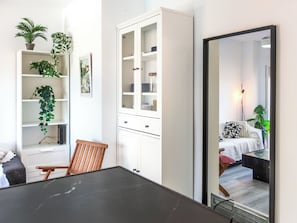 Quarto moderno com mesa de jantar, uma cadeira de madeira, armário com portas de vidro e um espelho grande refletindo uma cama com travesseiro estampado.