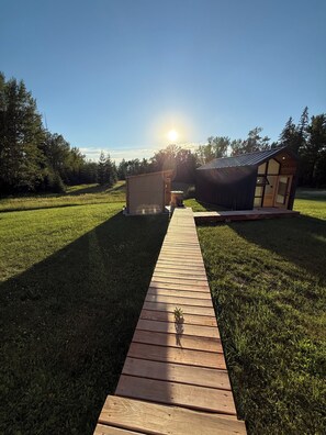 A wooden pathway leading to a small wooden structure with a glass door, surrounded by a grassy field and trees, under a clear sky.