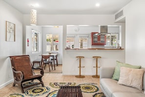 A living room with a sofa, a wooden chair, a patterned rug, a dining area with chairs, a kitchen with a counter and stools, and a wall-mounted clock.