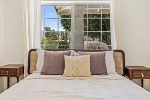 A bed with a white bedspread and multiple pillows, a window with a view of greenery, and wooden nightstands with drawers.