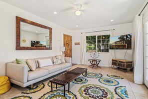A living room with a sofa, a coffee table, a wooden cabinet, a ceiling fan, a large mirror, and a window with a view of palm trees. - Resort-Style Desert Retreat with Huge Pool, Spa city id 069562 City ID # 069562 (Palm Springs)