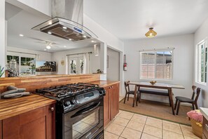 Een moderne keuken met een gasfornuis, oven en houten kastjes. Er is een eethoek met een tafel en stoelen.