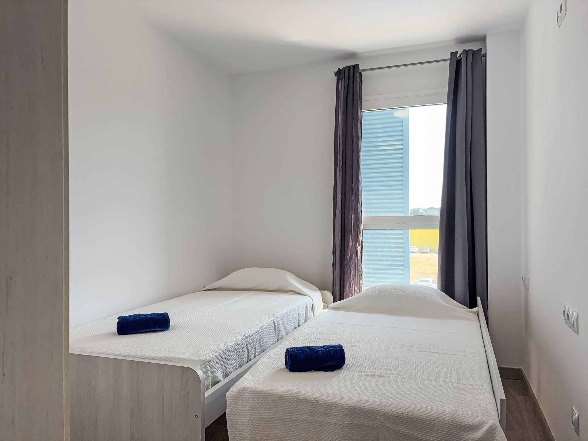 A small, clean room with two single beds, a window with curtains, and a view of the outside.
