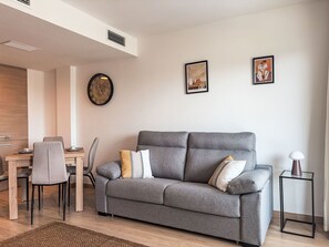 Ett modernt vardagsrum med en grå soffa, ett matbord med stolar, en klocka och två inramade tavlor på väggen.