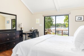 A bedroom with a large bed, a desk, a chair, a dresser, and a large window with a view of a dock and trees.