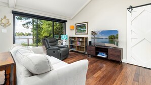 A spacious living room with a large window, a white sofa, a leather chair, a wooden coffee table, a bookshelf, and a TV on a wooden stand. - Lakefront 3-bedroom house on Poe Creek at Lake Gaston. Bring your boat! (Littleton)