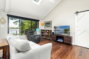 Ett rymligt vardagsrum med ett stort fönster, en vit soffa, en läderfåtölj, ett träbord, en bokhylla och en tv som står på ett träställ.