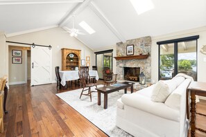 A spacious living room with a fireplace, a dining area with a wooden cabinet, and a large window offering a view of the outdoors.
