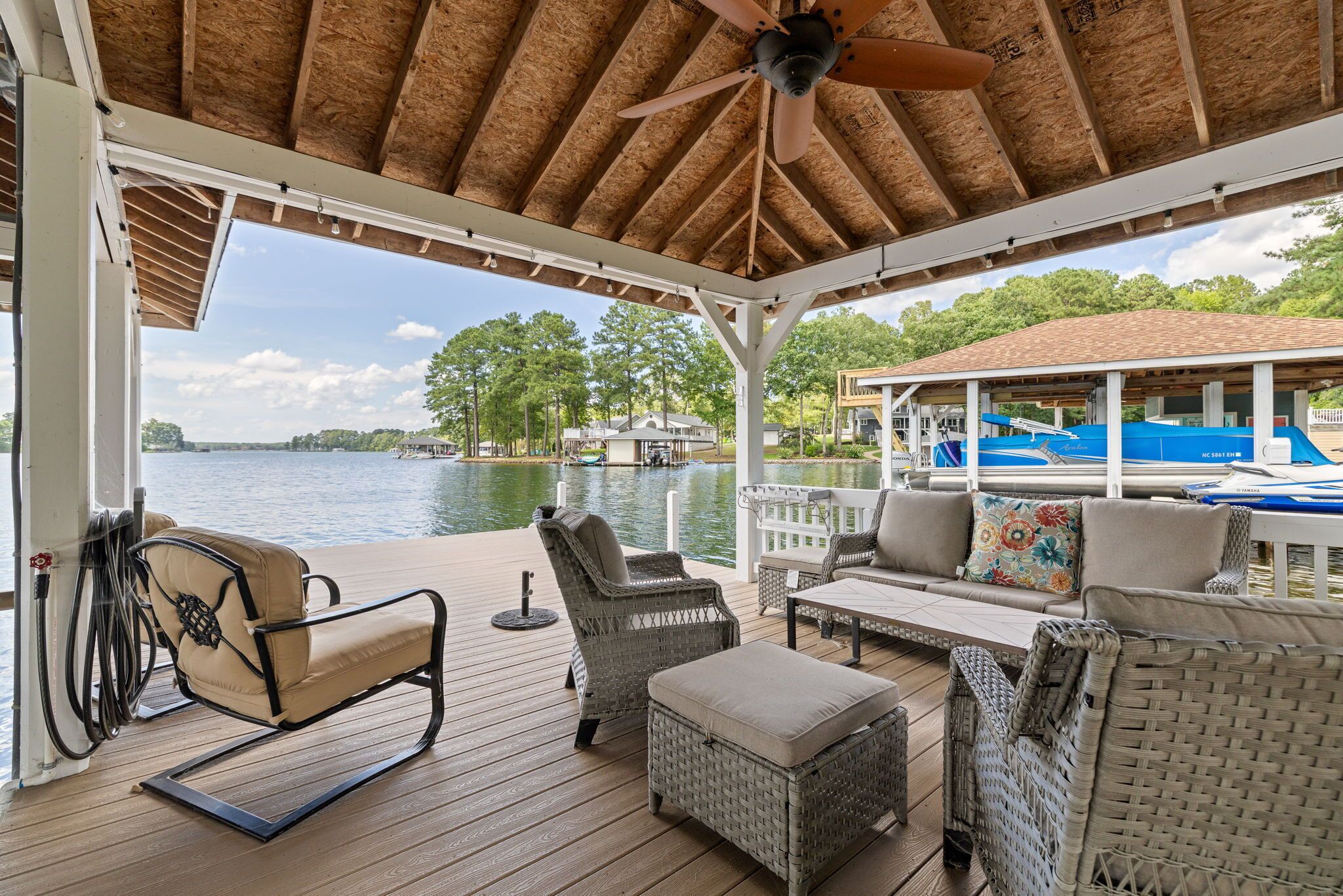 A covered outdoor deck with wicker furniture, a ceiling fan, and a view of a lake with boats.
