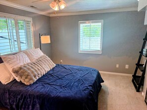 Ein Schlafzimmer mit einem Bett, zwei Fenstern mit Jalousien, einem Deckenventilator und einem Nachttisch.