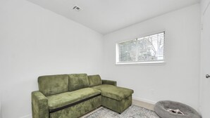 A green sectional sofa, a gray pet bed, a patterned rug, a window with blinds, and a white wall. - Chic & Cozy Home, Perfect for Work + Weekend stays, Near Main Highways (Houston)