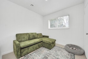 Ein grünes Ecksofa, ein graues Haustierbett, ein gemustertes Teppich und ein Fenster mit Jalousien, sowie eine weiße Wand.