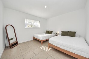 Ein Schlafzimmer mit zwei Betten, einem Fenster mit Jalousien, einem Nachttisch mit Lampe und einem Spiegel.