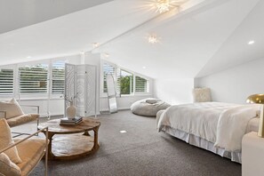 Une chambre moderne avec un plafond en pente, un lit, un coin salon avec une table et de grandes fenêtres équipées de stores.