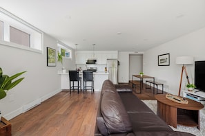 A modern living room with a brown leather sofa, a dining area with a wooden table, and a kitchen with white cabinets and stainless steel appliances. - Modern 3BR QE Park & 7-Min Walk to SkyTrain (Vancouver)