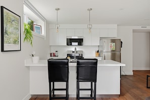A modern kitchen with white cabinetry, a central island with two black bar stools, a stainless steel refrigerator, and pendant lighting. - Modern 3BR QE Park & 7-Min Walk to SkyTrain (Vancouver)