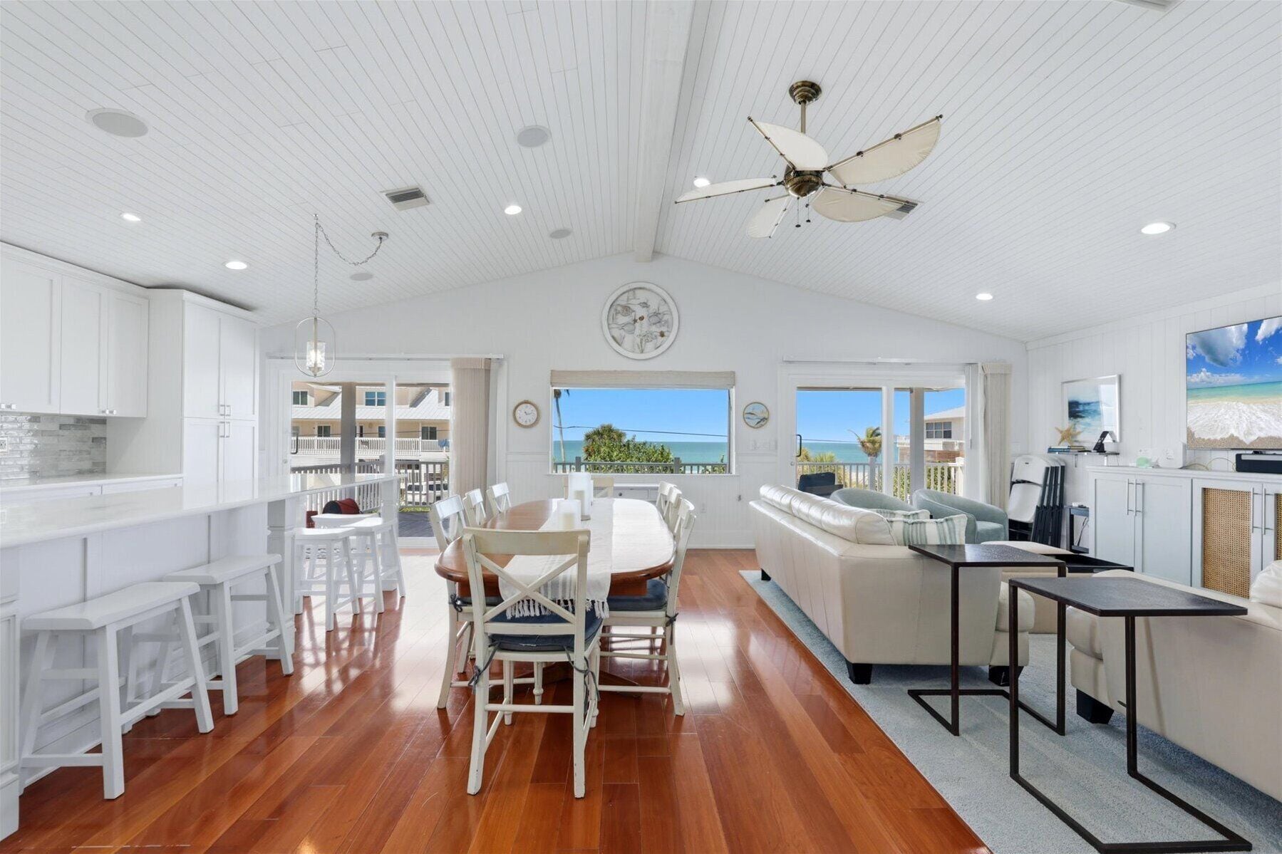 A spacious kitchen with white cabinetry, a dining area with a wooden table and chairs, a ceiling fan, and a view of the ocean.