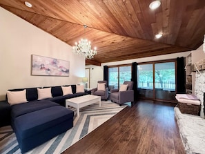 A modern living room with a wooden ceiling, a chandelier, a large sofa, armchairs, a coffee table, and a view through large windows. - Whimsy Wild: Walk to Downtown Saugatuck, Sauna, Hot Tub, Screen Porch, Fire Pit (Saugatuck)