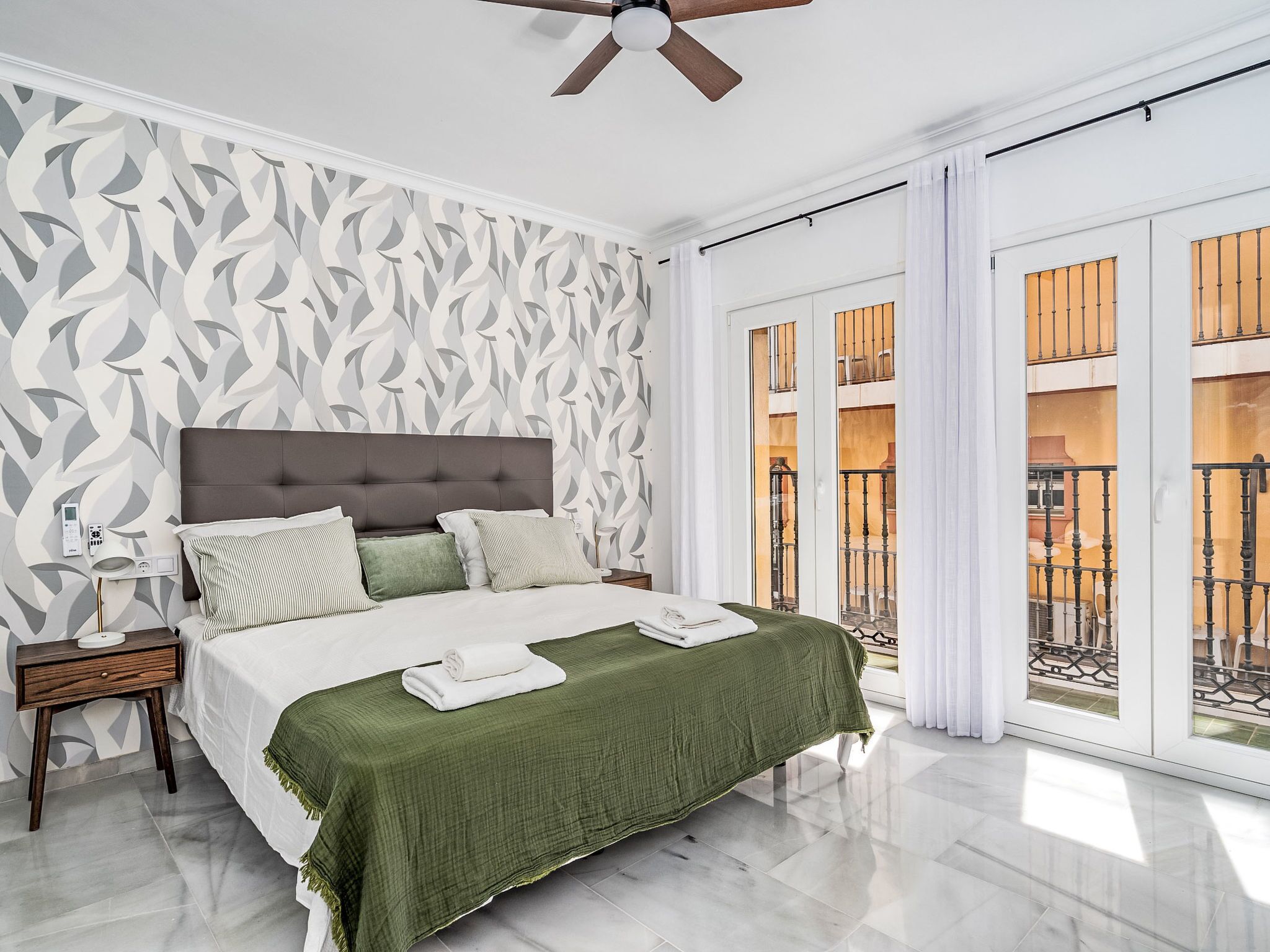 A bedroom with a large bed, a nightstand, a ceiling fan, and a view of the outside balcony.