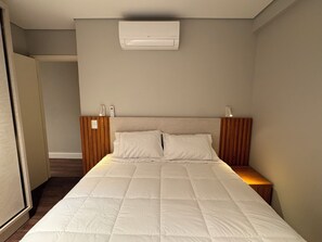 A bedroom with a bed, bedside tables, a wall-mounted air conditioner, and a door leading to another room.