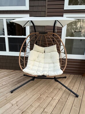Un fauteuil suspendu en osier, doté d’un siège et d’un dossier rembourrés, soutenu par des pieds en métal, et placé sur une terrasse en bois.