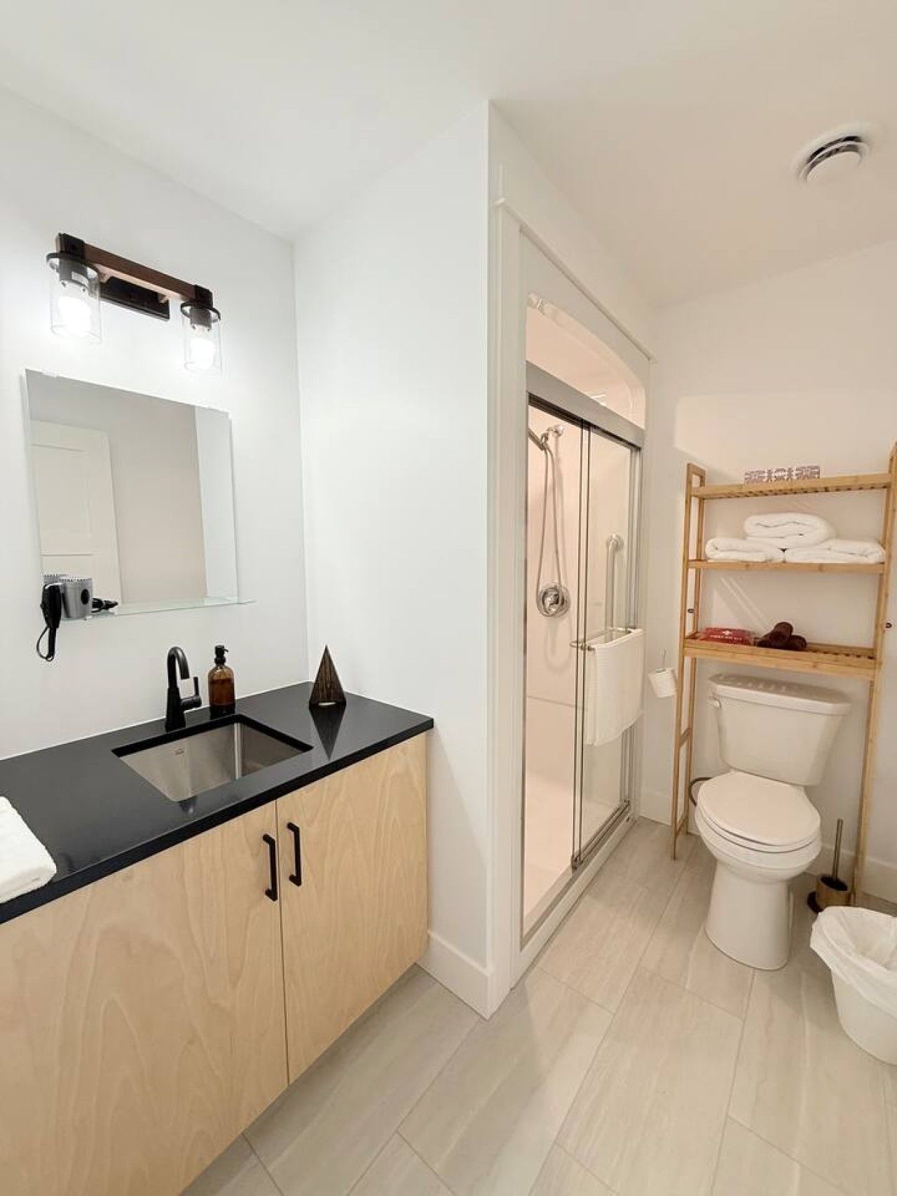 A modern bathroom with a black countertop sink, a mirror, a shower enclosure, and a toilet with a shelf above it.