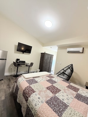 A bedroom with a bed, a desk, a chair, a TV, and an air conditioning unit.