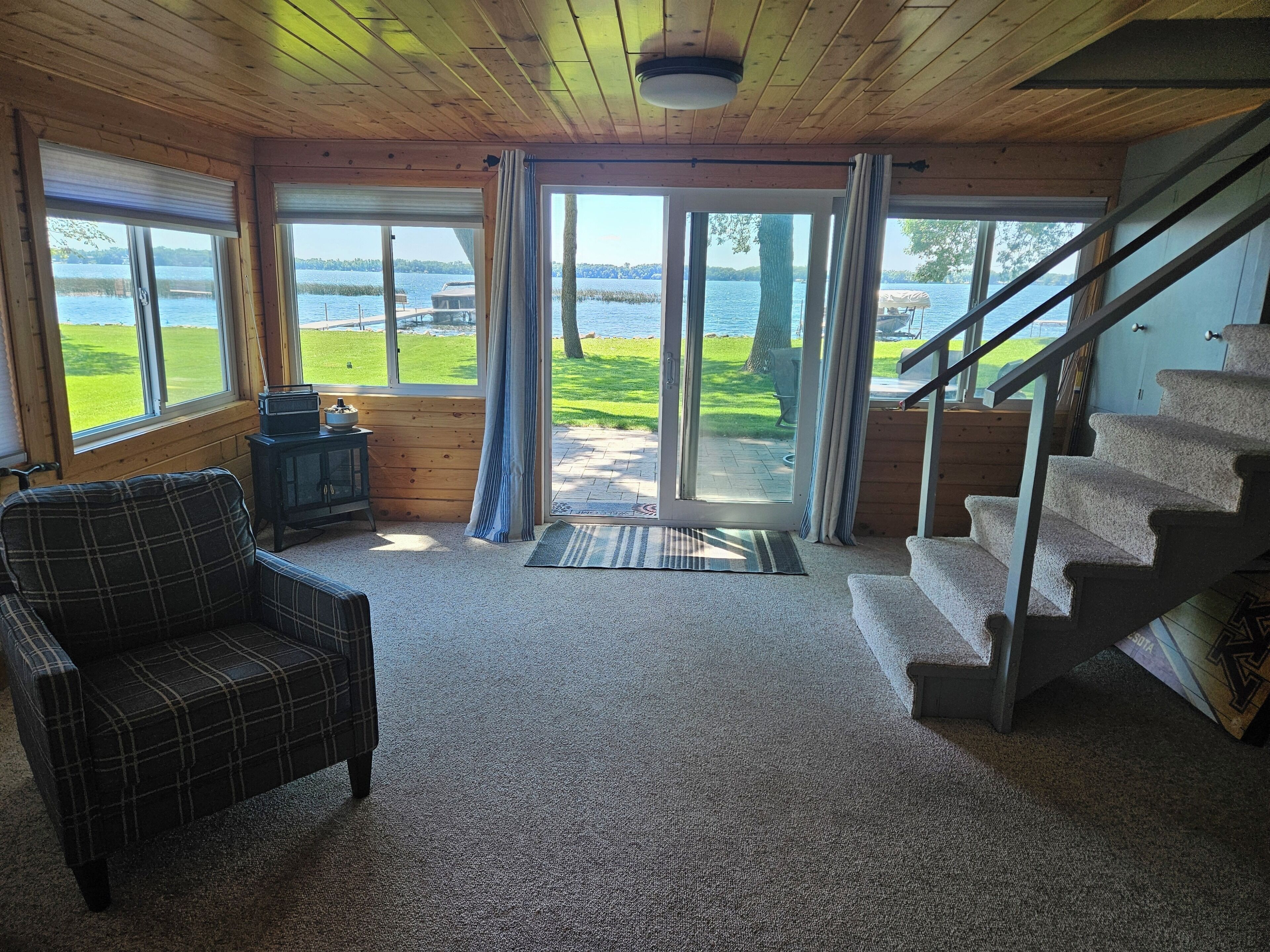 A cabin interior with a plaid chair, a small table, a fireplace, and a staircase leading to a deck with a view of a lake.