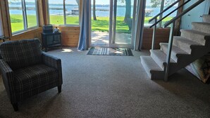 A cabin interior with a plaid chair, a small table, a fireplace, and a staircase leading to a deck with a view of a lake.