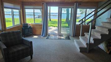 A cabin interior with a plaid chair, a small table, a fireplace, and a staircase leading to a deck with a view of a lake.