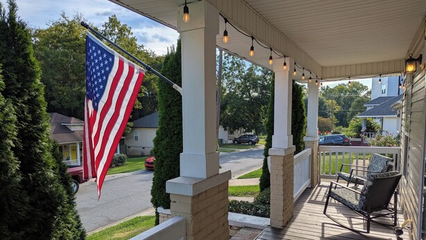 Eine überdachte Veranda mit einer Flagge, Sitzgelegenheiten im Freien und Blick auf eine Straße.
