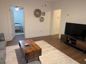 Ein Wohnzimmer mit Couchtisch aus Holz, einem Fernseher auf einem Holzgestell und einer Tür, die in ein Schlafzimmer führt.