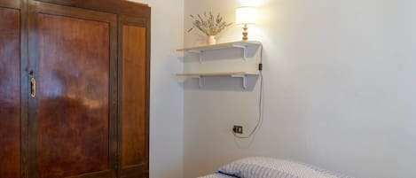 A bedroom with a wooden wardrobe, a bed with patterned bedding, a wall-mounted shelf with a lamp, and a ceiling with wooden beams.