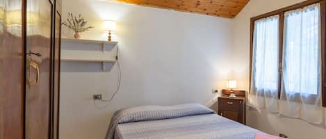 Un dormitorio con techo de madera, una cama con ropa de cama estampada, una mesita de noche con lámpara y una ventana con cortinas translúcidas.