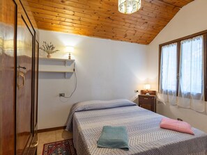 Ein Schlafzimmer mit Holzbalkendecke, einem Bett mit gemusterten Betttextilien, einem Nachttisch mit Lampe und einem Fenster mit durchsichtigen Vorhängen.