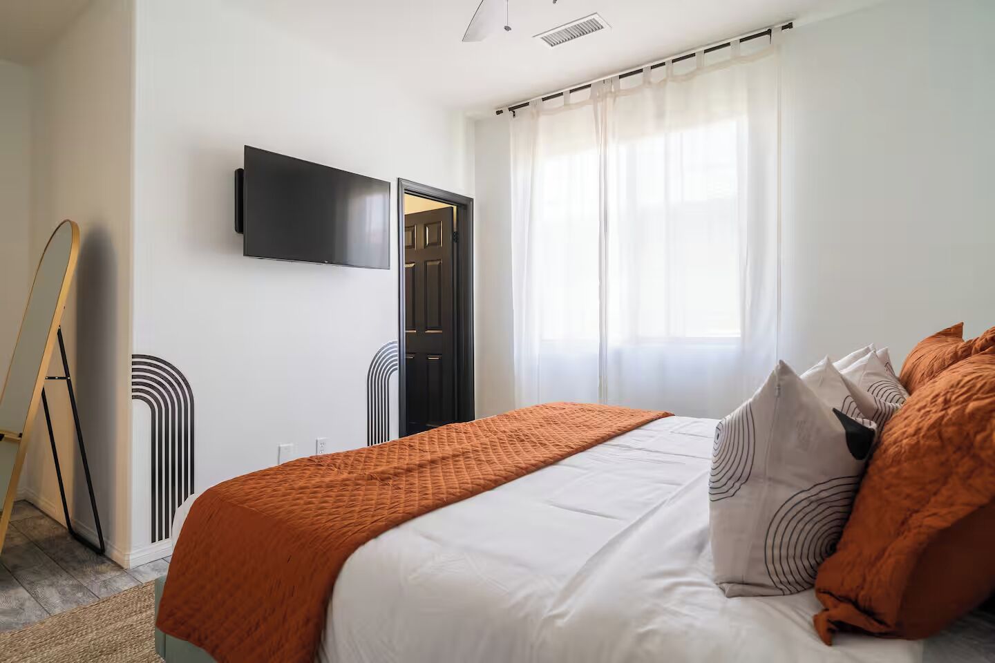 A bedroom with a bed, a television mounted on the wall, a door, and a window with curtains.