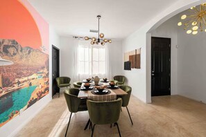 A dining area with a table set for four, green chairs, a chandelier, and a large painting on the wall. - Golden Hour - Pool, Spa, BBQ, Mini-Golf, Gameroom! (Indio)
