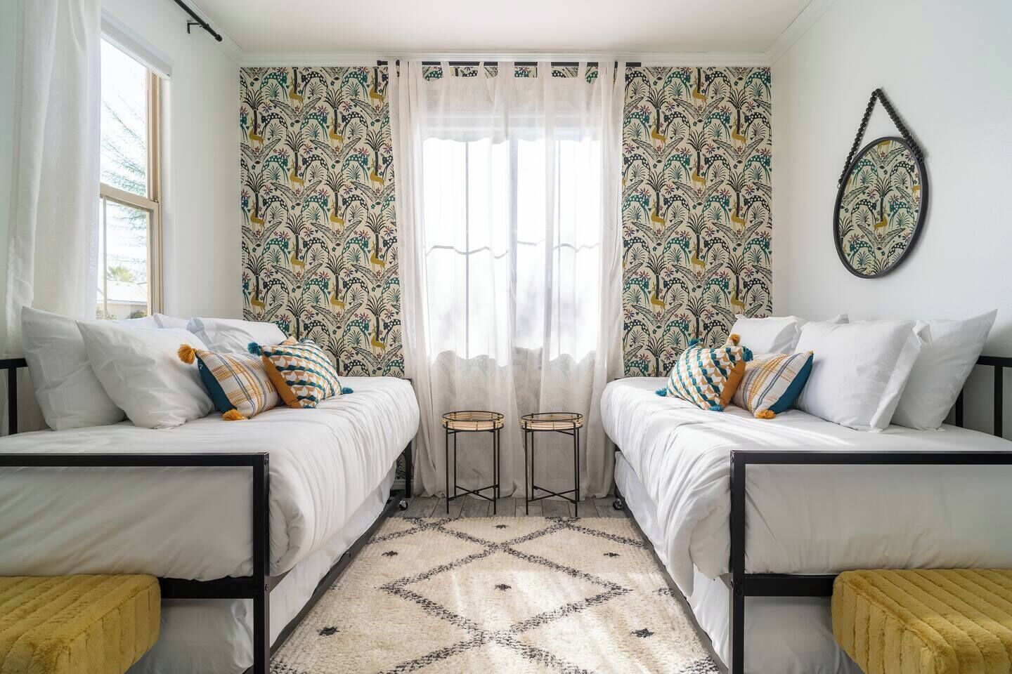Quarto com duas camas, janela com cortinas leves, papel de parede estampado e uma pequena mesa redonda.